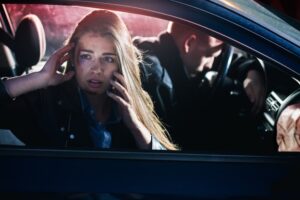 A distressed woman sits in the passenger seat, reaching out for help as she dials the emergency hotline.