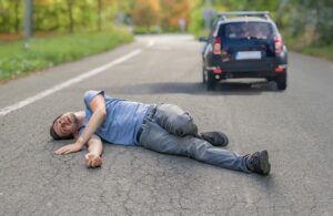 A hit-and-run scenario unfolds as a man lies injured on the road, directly in front of an idling car.
