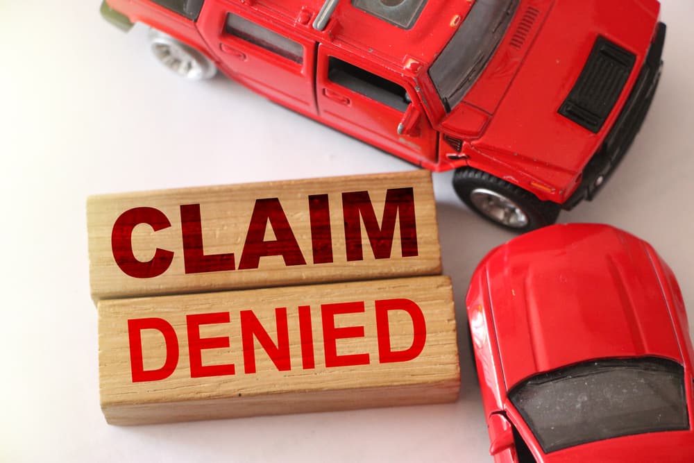Wooden blocks spell out "Claim denied" while two red cars are shown in a road accident on a white background. This represents the concept of insurance denial, indicating a negative response from the insurance company.