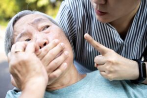 An angry Asian woman covers a senior's mouth while making a silence gesture, symbolizing fear, social violence, and the aggression that may silence vulnerable elderly individuals.