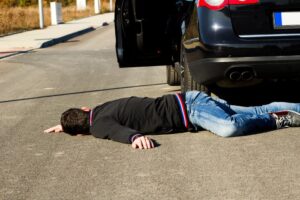 A man lies on the road behind a black car after a collision. The pedestrian, struck by the vehicle, is on the ground.