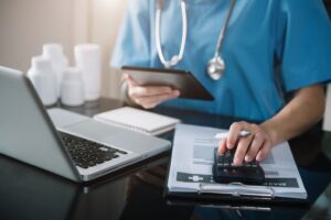 A doctor uses a tablet and calculator at the clinic to determine medical fees, costs, and revenue, focusing on accurate billing and financial management.