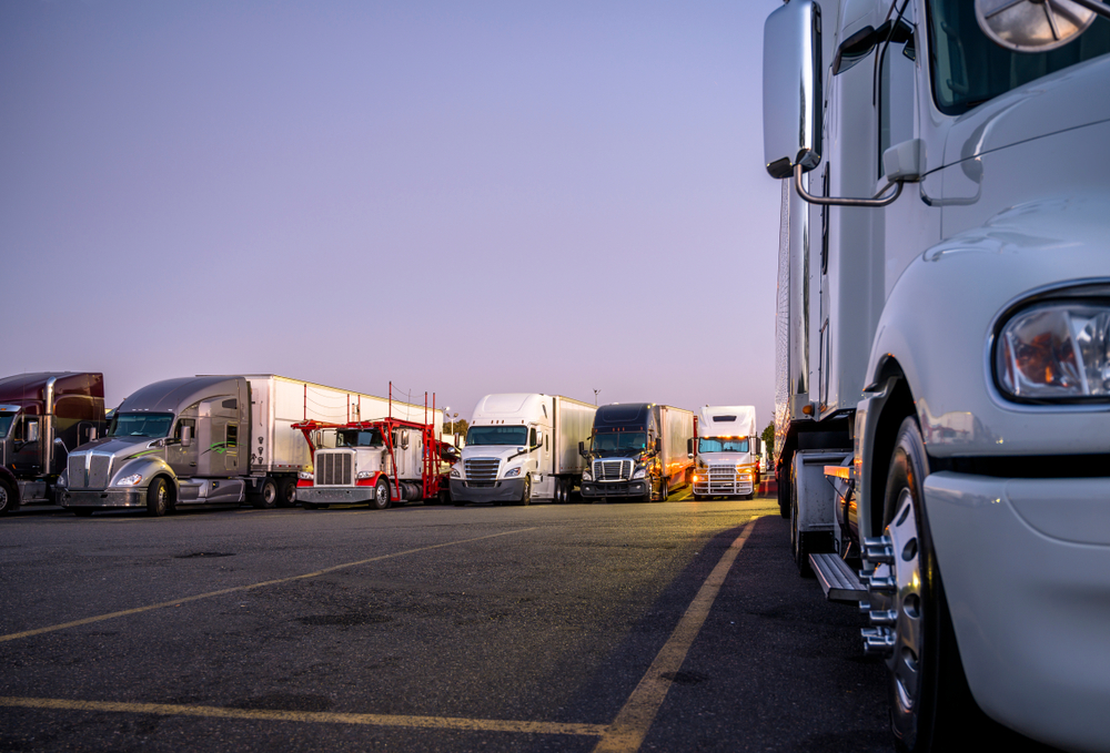 Different make and models big rigs semi trucks with semi trailers standing in row on truck stop parking lot for rest and comply with the movement according to the schedule for successful delivery
